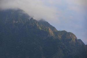 mörk atmosfärisk overkligt landskap med en mörk klippig berg topp i låg moln i en grå molnig himmel. en grå låg moln på en hög topp. hög svart sten i låg moln. overkligt dyster berg. foto