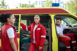 paramedics och läkare stående på de sida ambulans. läkare är bärande en medicinsk trauma väska. grupp av tre paramedics stående i främre av ambulans med leende. foto