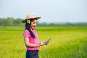asiatisk kvinna jordbrukare innehav läsplatta gående i ris fält till Lagra information foto