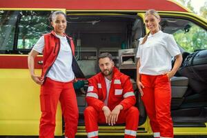 en multietnisk grupp av tre paramedics på de bak- av ett ambulans, klättrande i genom de öppen dörrar. de två kvinnor är leende på de kamera, och deras manlig kollega har en allvarlig uttryck. foto