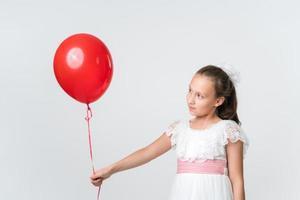 Söt flicka i vit klänning innehav röd ballong i utsträckt hand, ser eftertänksamt på ballong foto