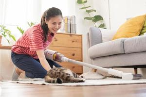 Lycklig asiatisk ung hushållerska kvinna använder sig av Vakuum rengöring, rengöringsmedel till ta bort damm, hår eller päls på golv i levande rum medan söt katt liggande på matta. rutin- hushållsarbete, Syssla i hushåll av hembiträde. foto