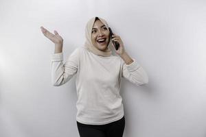 upphetsad asiatisk kvinna bär hijab pekande på de kopia Plats bredvid henne medan innehav henne telefon, isolerat förbi vit bakgrund foto