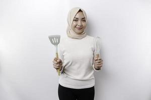 upphetsad asiatisk muslim kvinna bär en huvudduk, innehav matlagning gods och leende, isolerat förbi vit bakgrund foto