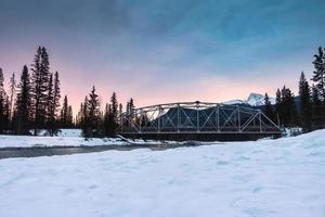 soluppgång över snöig klippig muntains och de bro korsa de flod i tall skog på vinter- på banff nationell parkera foto