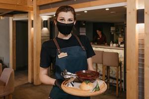 en servitris som bär en svart ansiktsmask och engångshandskar serverar kunder på ett café foto