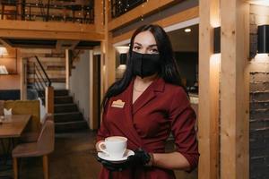 en kvinnlig restaurangchef bär en svart ansiktsmask och engångshandskar som håller en kopp kaffe i en restaurang