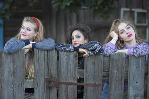 tre rolig by flickor är stående nära de gammal by staket. uttråkad Land flickor. foto