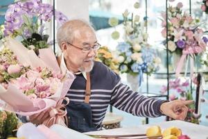 e-handel företag, ett senior manlig blomsterhandlare arbetare demonstrera och visa blommig arrangemang via uppkopplad leva strömning med smartphone Ansökan i en ljus blomma affär, en skön blommar Lagra. foto