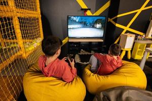 två bröder spelar lopp video spel trösta, Sammanträde på gul pouf i barn spela Centrum. foto