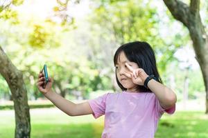 skön asiatisk barn flicka använder sig av mobil smartphone. liten flicka är spelar i digital spel i de grön parkera. foto