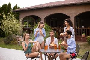 grupp av ung människor glädjande med cider och äter pizza förbi de slå samman i de trädgård foto