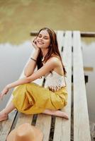en hippie kvinna sitter med henne hatt på en bro förbi en sjö med henne händer upp i de luft på en natur resa och leende lyckligt i eko-kläder. avslappnad livsstil foto