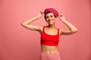 ung atletisk kvinna med en kort frisyr och lila hår i en röd topp och en rosa hatt med ett atletisk figur ler och grimaser som visar tecken med henne händer ser på de kamera på en rosa bakgrund foto