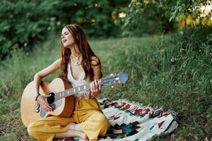 ung kvinna hippie konstnär spelar gitarr och sjunger låtar i miljövänlig Kläder Sammanträde på de jord utanför i natur i de höst ser ut på de solnedgång foto