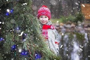 en barn på jul gata. en pojke i en röd stickat hatt står nära en dekorerad jul träd.a pojke på en vinter- promenad. foto