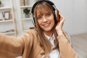 söt ung blond studerande lady frilansare i värma Tröja i hörlurar lyssna fav låtar Sammanträde i fåtölj på modern Hem interiör. musik tid avkopplande Häftigt Spellista begrepp. bred selfie se foto