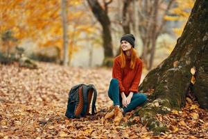 kvinna i byxor och stövlar med en ryggsäck sitter nära en träd i de höst skog fallen löv foto