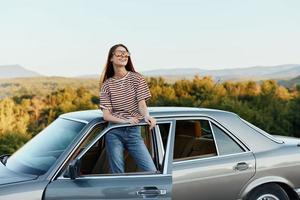 en ung kvinna förare utseende ut av de bil på de höst landskap och ler tillfredsställande foto
