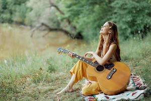 hippie kvinna eco vänlig spelar gitarr ler och sång låtar i natur Sammanträde på en pläd förbi de sjö i de kväll i de solnedgång solljus. en livsstil i harmoni med de kropp och natur foto