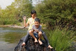 asiatisk flickor och Pojkar ta en ha sönder från spelar i de flod till sitta på de rocks. både av dem skrattade och undvek de vatten den där hade varit hällde över dem. vatten droppar spridning och se färsk. foto