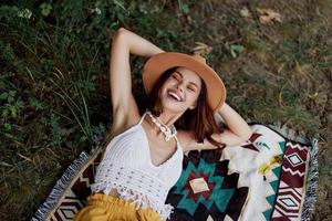 en kvinna i miljövänlig Kläder i en hippie se är liggande på en färgad pläd leende och ser på ett höst solnedgång i natur i de parkera. livsstil på en resa av harmoni med de värld foto