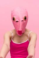 en kvinna bär en silikon halloween mask i de form av en rosa fisk med stor gul ögon utseende på de kamera mot en rosa bakgrund foto