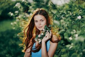 en kvinna med flygande hår i vår står nära en blomning äpple träd och ler ser på de kamera i en blå klänning, lycka foto