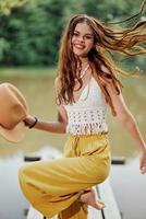 en ung kvinna i en hippie se och eko-klänning dans utomhus förbi de sjö bär en hatt och gul byxor i de sommar solnedgång foto