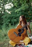 en flicka i eko-kläder hippie Sammanträde med en gitarr och ser på en solnedgång i de sommar foto