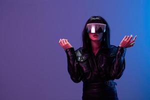 förvirrad chockade Söt brunett lady i läder jacka trendig spekulär solglasögon öppen mun spridning händer Framställ isolerat i blå violett Färg ljus bakgrund. neon fest cyberpunk begrepp. kopia Plats foto