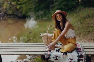 Lycklig flicka sitter i eco kläder på en bro förbi de sjö bär en hatt i en hippie se och leende foto