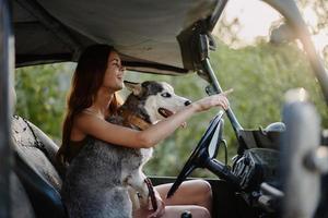 en skön ung kvinna sitter Bakom de hjul av henne bil tillsammans med en hes ras hund och ler glatt åtnjuter de resa foto