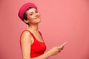 ung kvinna bloggare med färgad rosa hår och kort frisyr vända genom de skärm av de telefon och skriver en meddelande med en leende i eleganta kläder på en rosa bakgrund foto