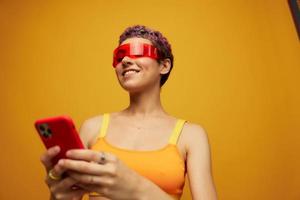 kvinna bloggare leende i ovanlig röd tusenåriga glasögon innehav henne telefon och ser på de skärm mot ett orange bakgrund i en eleganta gul topp foto