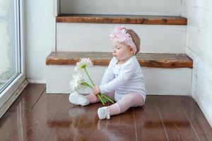 liten bebis flicka bär vår krans lokaliserings på golv i ljus ljus levande rum nära fönster och spelar med gerbera blommor foto