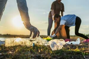 människor volontär- förvaring sopor plast flaska in i svart väska på parkera nära flod i solnedgång foto