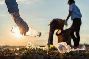 människor volontär- förvaring sopor plast flaska in i svart väska på parkera flod i solnedgång foto