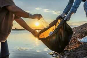 människor volontär- förvaring sopor plast flaska in i svart väska på flod i solnedgång foto