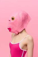 en kvinna i en rosa fisk huvud mask står i profil mot en rosa bakgrund och utseende in i de kamera med ett gul öga, en galen konceptuell halloween kostym foto