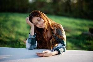 flicka med telefon i hand Sammanträde i natur i de parkera på de tabell leende vackert och ser på de kamera med henne röd hår belyst förbi de solnedgång solljus av sommar foto