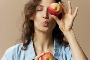 rolig söt lockigt kvinna i jeans tillfällig skjorta stänga öga med äpple skickar kyss på kamera Framställ isolerat på över beige pastell bakgrund. friska mat. naturlig miljövänlig Produkter begrepp. kopia Plats foto