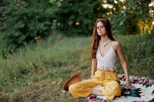 en ung hippie kvinna mediterar i natur i de parkera, Sammanträde i en lotus placera på henne färgrik pläd och njuter harmoni med de värld i eko-kläder foto