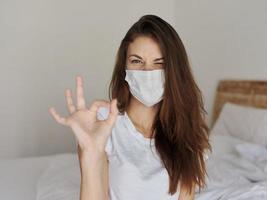 kvinna på Sammanträde på säng bär medicinsk mask positiv hand gest foto