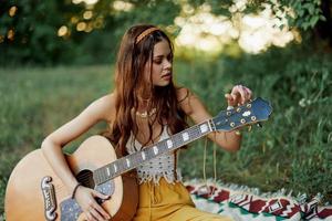 flicka hippie kvinna spelar gitarr i miljövänlig kläder Sammanträde på de jord utanför i natur i de falla tittar på de solnedgång foto