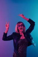 haft sexig skön blond kvinna i läder jacka glittrande klänning trendig solglasögon dansa håll händer upp visa v-tecken Framställ isolerat i blå rosa Färg ljus studio bakgrund. neon fest begrepp foto