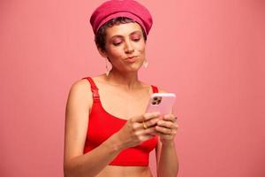 ung kvinna bloggare med färgad rosa hår och kort frisyr vända genom de telefon skärm och skriver en meddelande med en leende och överraskning i eleganta kläder på en rosa bakgrund foto