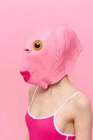 en kvinna i en rosa fisk huvud mask står i profil mot en rosa bakgrund och utseende in i de kamera med ett gul öga, en galen konceptuell halloween kostym foto