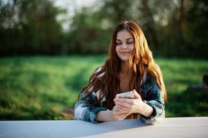 en skön kvinna avkopplande och arbetssätt på henne telefon Sammanträde i natur i de parkera bland de träd leende och innehav henne smartphone i henne hand belyst förbi de ljus solnedgång ljus foto