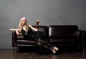 skön blond kvinna på de soffa med en flaska av öl och i en svart klänning foto